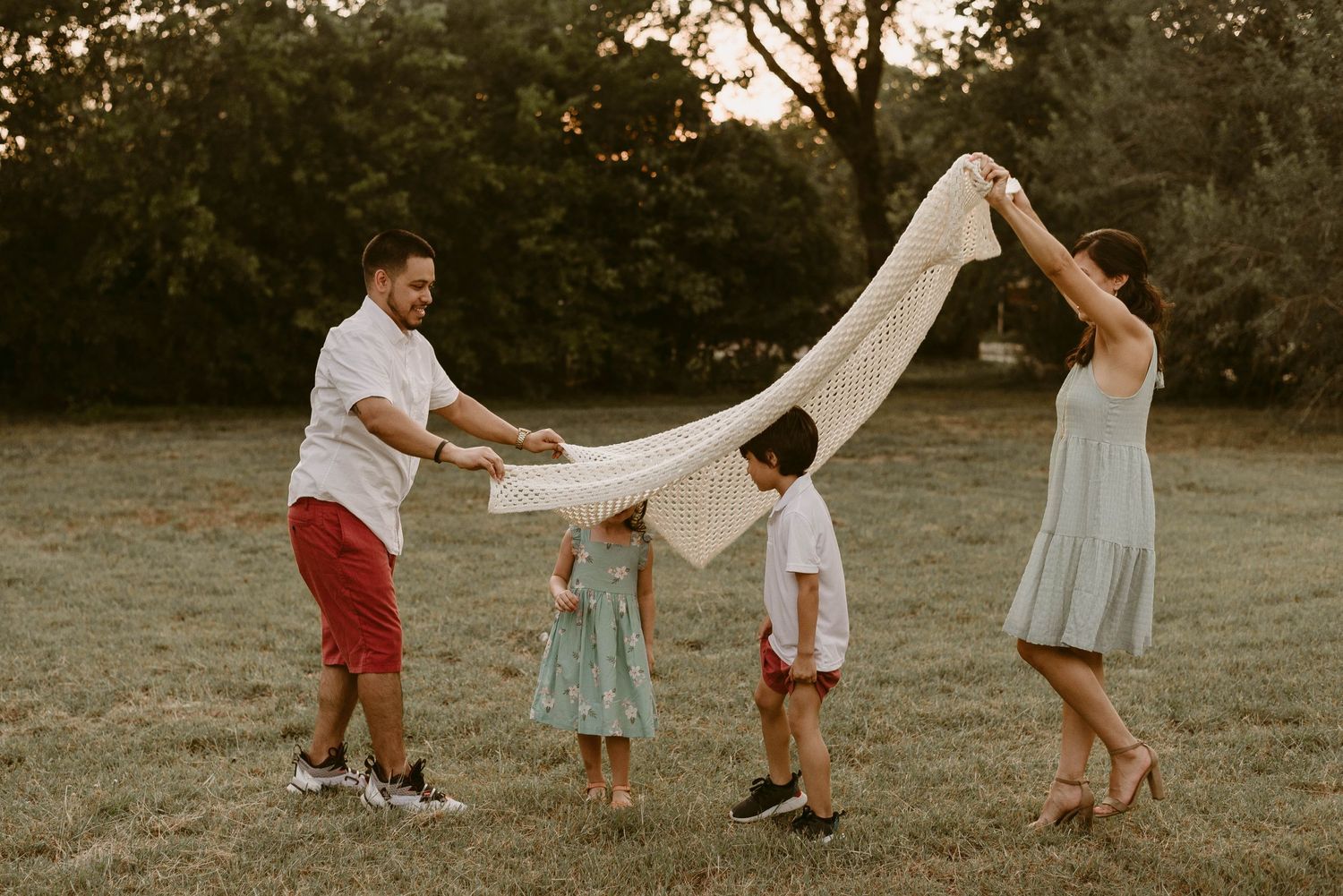 Family plays together outdoors holding white fabric above their heads in summer field.
