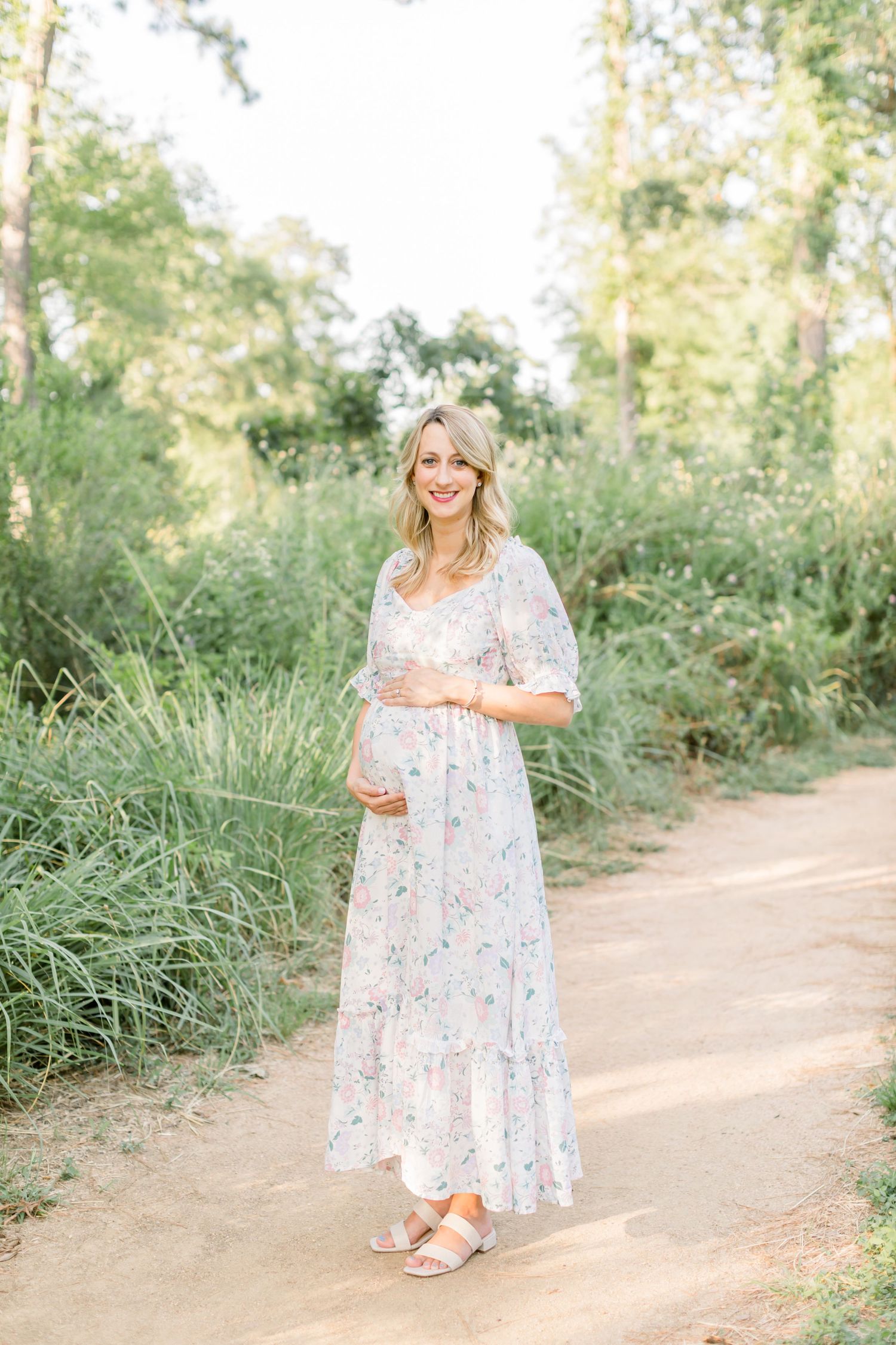 An expectant mother in a floral maxi dress poses on a nature trail.
