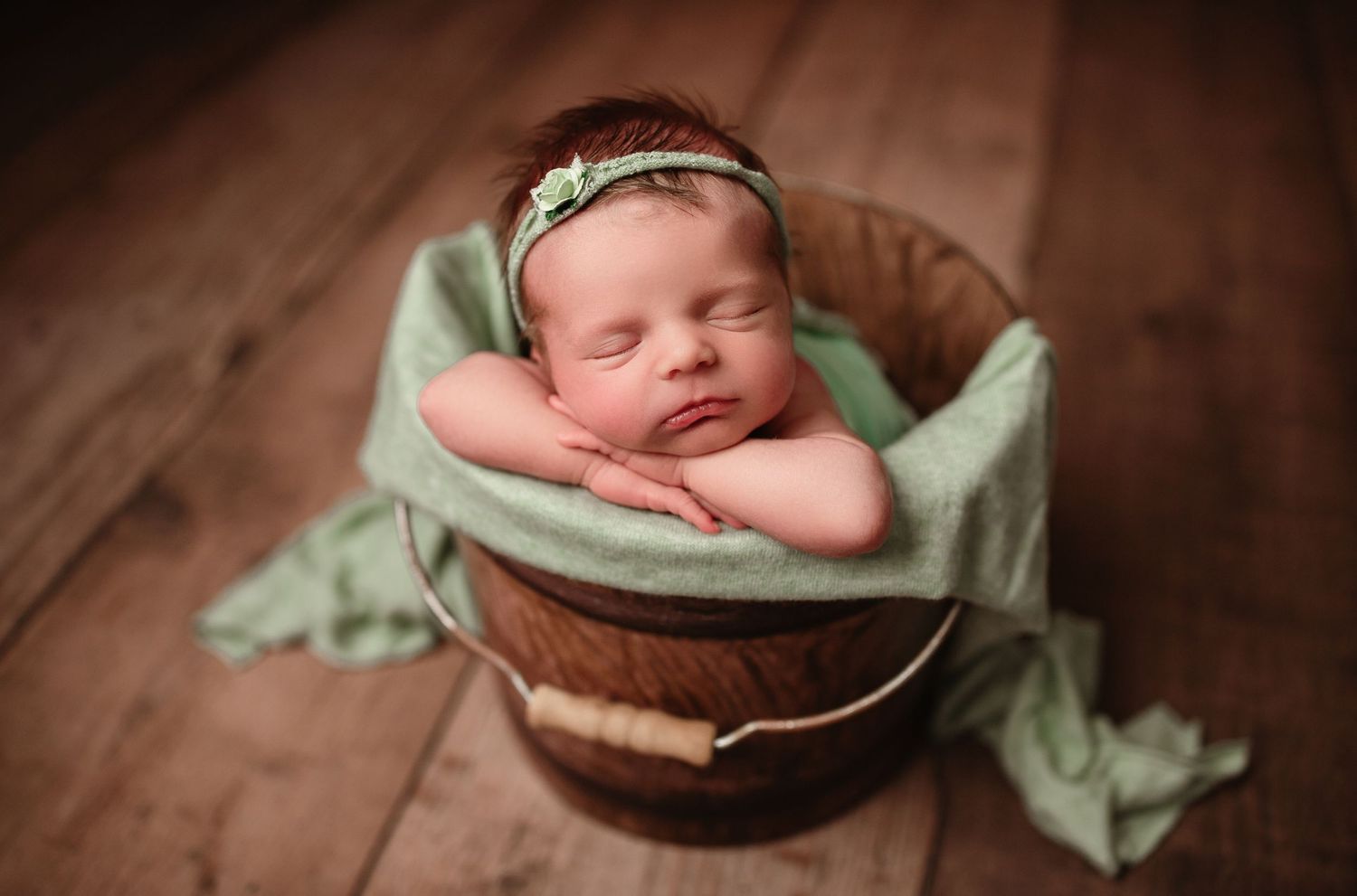 Newborn sleeping peacefully in mint green wrap inside rustic wooden bucket.