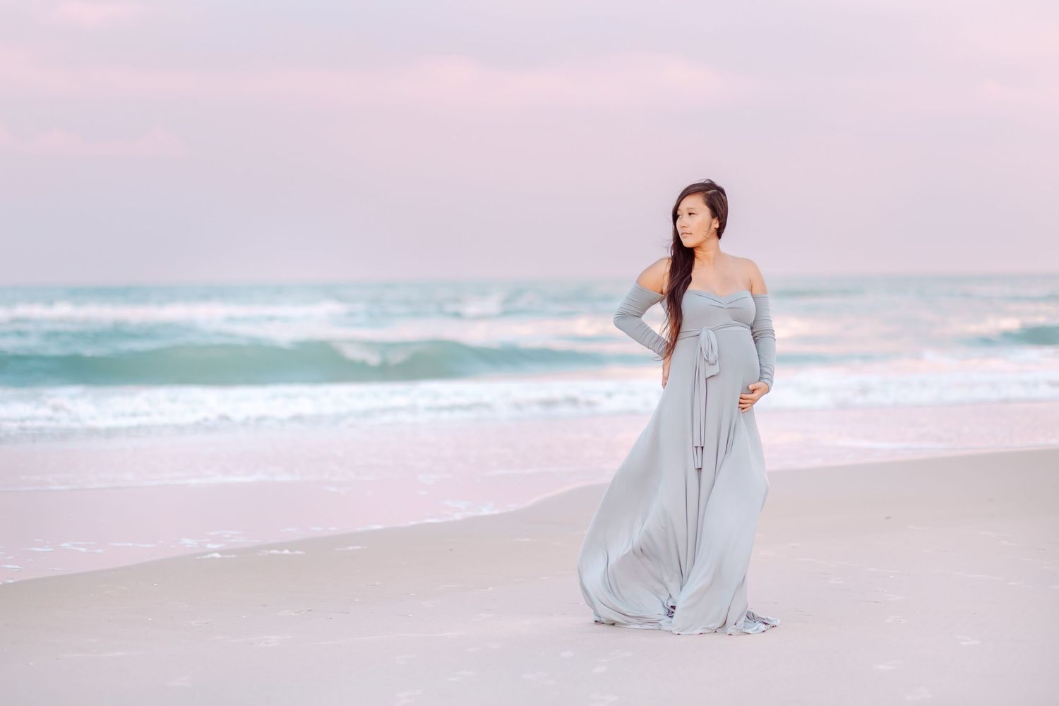 Woman in off-shoulder grey gown poses on pink sandy beach at sunset.