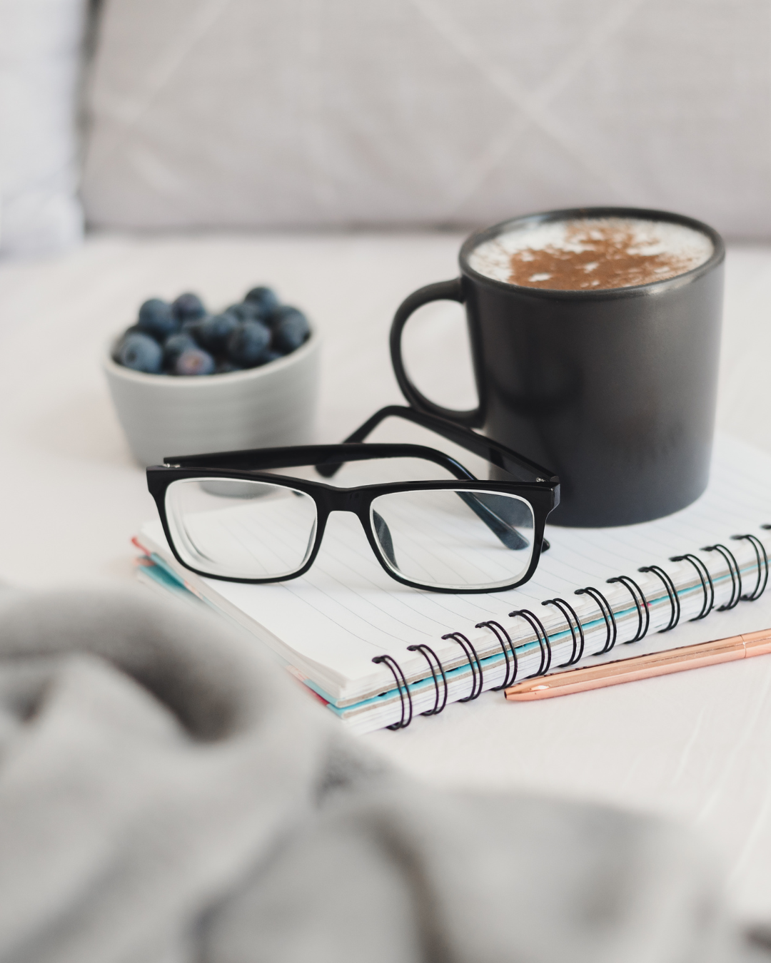 Workspace arrangement with notebook, glasses, coffee cup and blueberries on white surface.  L Visions Photography.