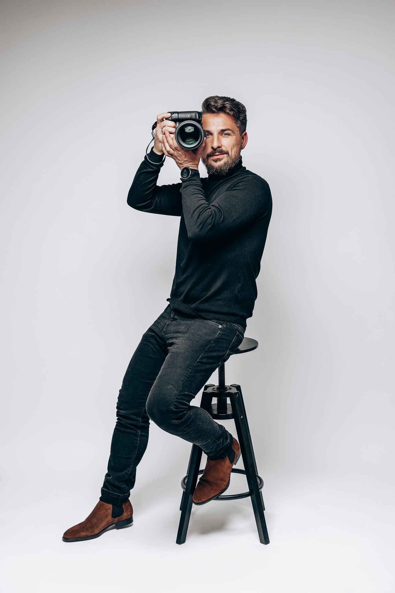Professional photographer in black sweater posing with camera on stool against white background.