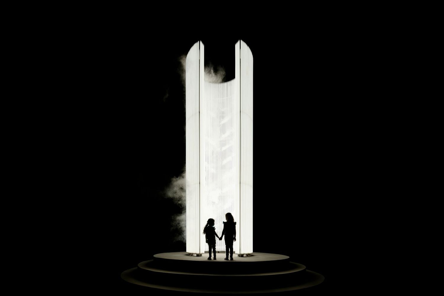 Silhouette of two people standing between tall illuminated pillars in a dramatic black and white composition.