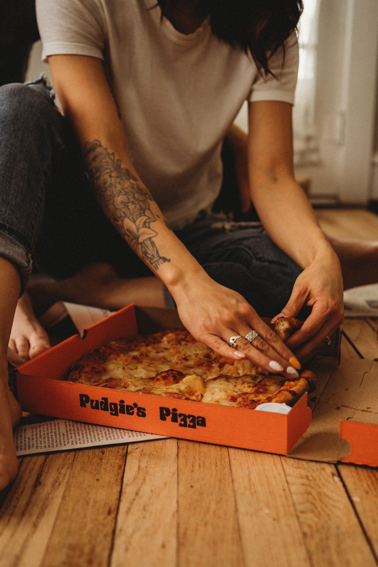 Tattooed hand reaching for slice of Pudgie's pizza on wooden floor with newspaper.