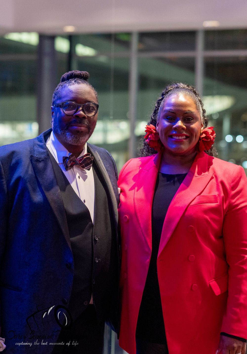 Two people pose together wearing contrasting black and red formal suits.
