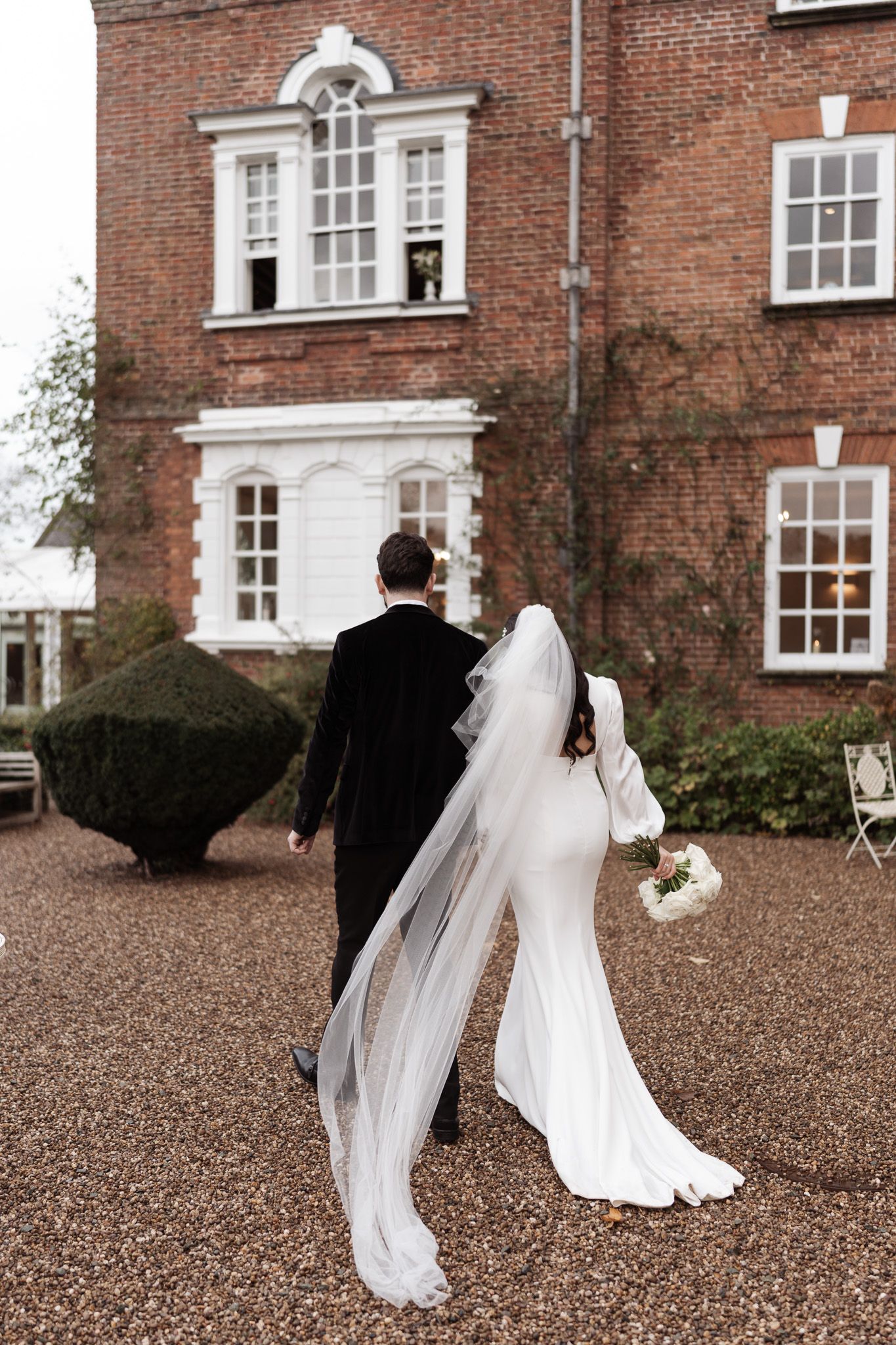 Wedding couple walking along gravel path outside historic brick building with flowing veil.