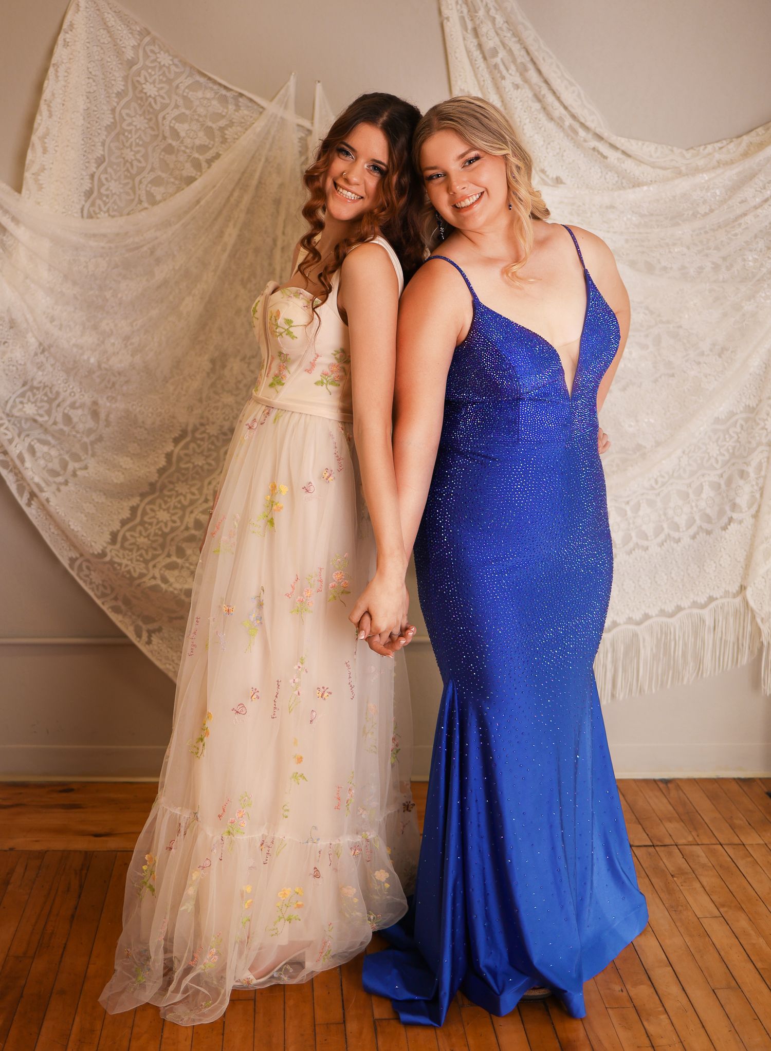 Two formal dresses - one in cream tulle with gold details and one in royal blue - against a lace backdrop.
