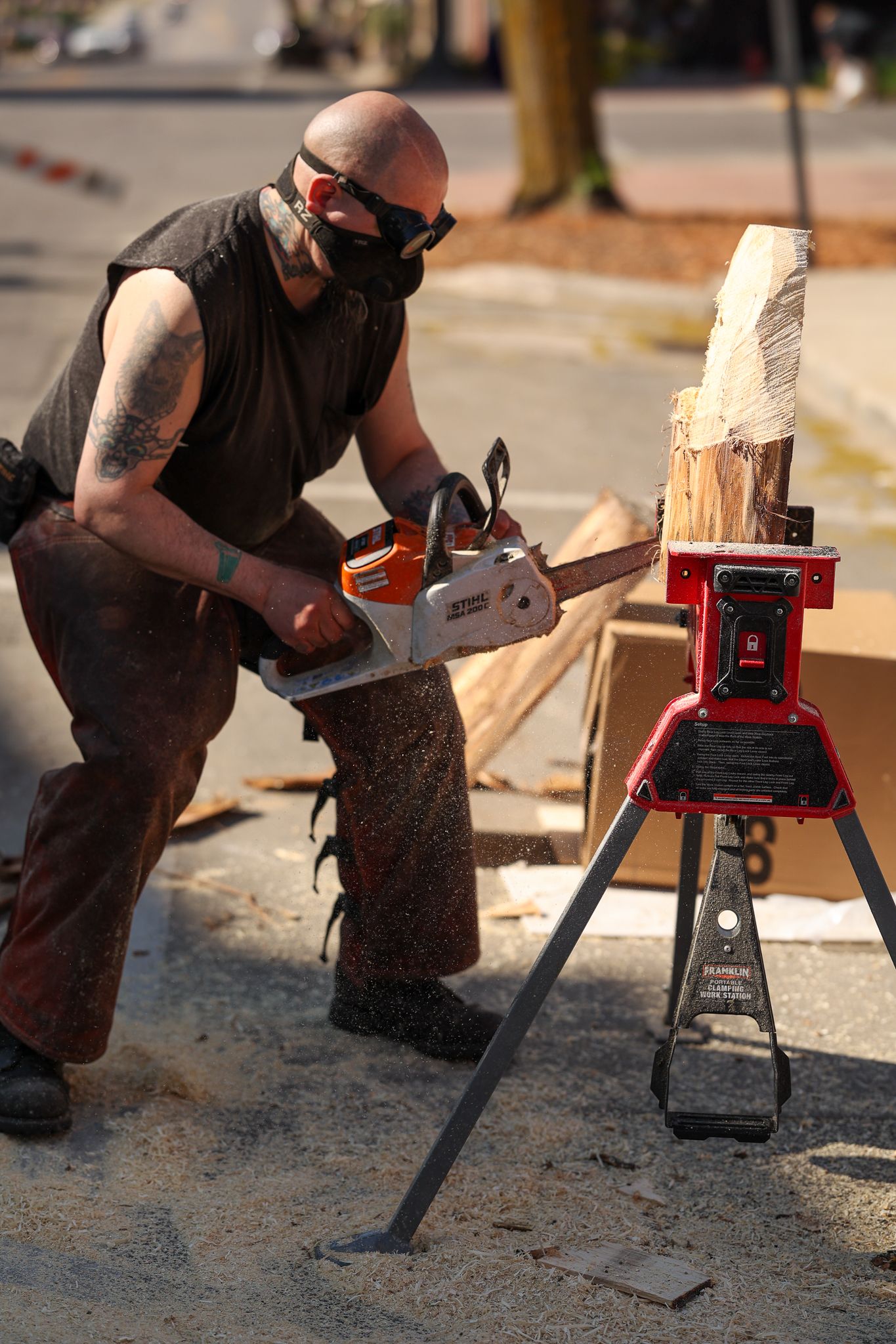 Craftsman using chainsaw on wooden log with safety equipment and work stand.