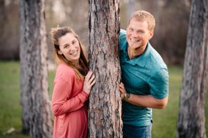A playful couple peeking around opposite sides of a pine tree trunk.