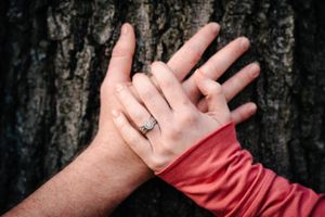 Close-up of intertwined hands with engagement ring against dark tree bark.