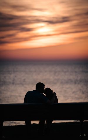 Silhouette of romantic couple on bench against dramatic orange sunset over water.