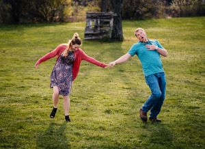 Two people dance playfully in a grassy field with a rustic backdrop.