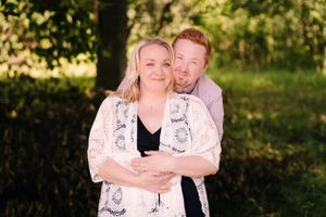 A couple embraces while standing in a sunlit outdoor setting with trees in the background.