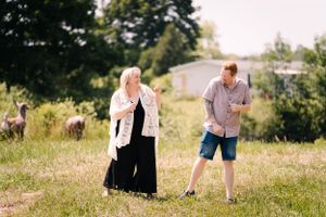 Two people stand in a grassy field with trees and buildings in the background.