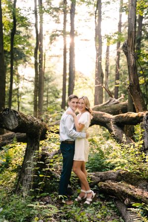 A couple embraces in a sun-dappled forest with fallen logs and golden light filtering through trees.