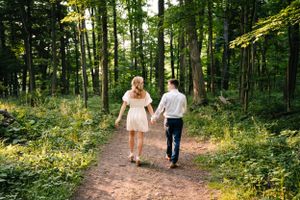 A couple in light clothing walks hand in hand down a forest path surrounded by lush greenery.