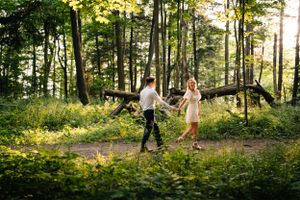 A couple dances playfully on a forest path with sunlight streaming through the trees.