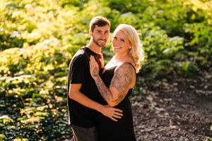 A couple embraces while posing in front of lush green foliage on a sunny day.