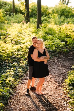 A couple embraces on a sunlit forest path surrounded by lush greenery and golden light.