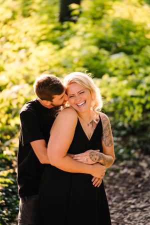 An intimate moment captured between a couple sharing laughter in a sun-drenched wooded area.