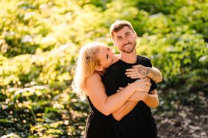 A playful embrace between two people wearing black against a backdrop of sunlit foliage.