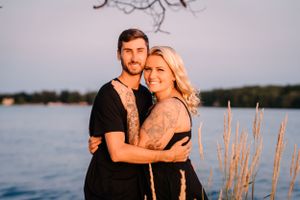 A couple stands close together at sunset near a waterfront with cattails in the background.