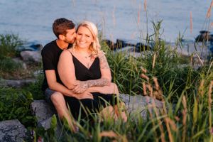 A couple sits together on rocky shoreline with water and tall grasses in the background.
