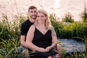 Couple embraces at sunset by a waterfront with tall grass and natural lighting.