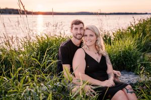 Romantic sunset photoshoot with couple sitting by lakeside surrounded by tall grass.