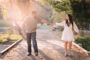 A couple holds hands while walking on railroad tracks at sunset with golden light streaming through trees.