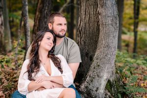 A couple sits against a tree trunk in a serene forest setting during autumn.