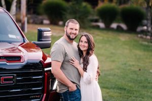 A couple poses together beside a red GMC truck on a grassy lawn.