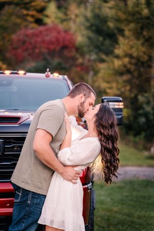 A romantic couple shares a tender kiss in front of a red pickup truck with autumn foliage in the background.