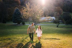 A couple walks hand in hand through a sunlit meadow with mountains in the background.