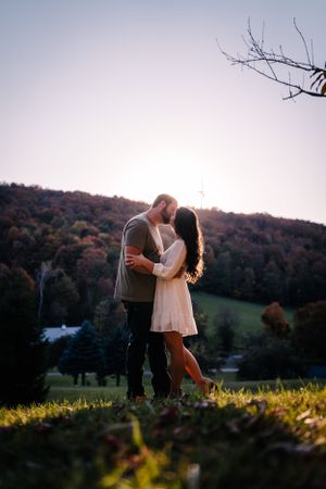 A couple shares an intimate moment at sunset with rolling hills and autumn foliage in the background.