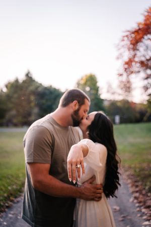 A couple shares a tender moment outdoors while showing off an engagement ring during autumn.