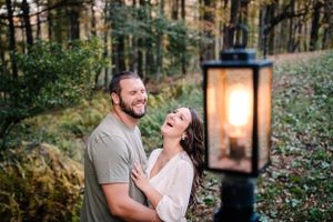 A couple laughs together next to a glowing vintage lantern in a wooded setting.