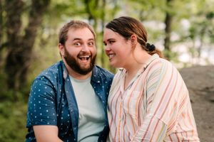 A couple laughs together during an outdoor photo session in a wooded setting.