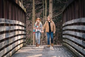 Couple walking on a wooden bridge surrounded by tall trees in a forest setting.