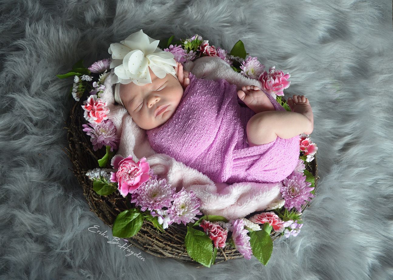 Newborn baby sleeping in a floral nest wrapped in pink with a white bow headband on gray fur.