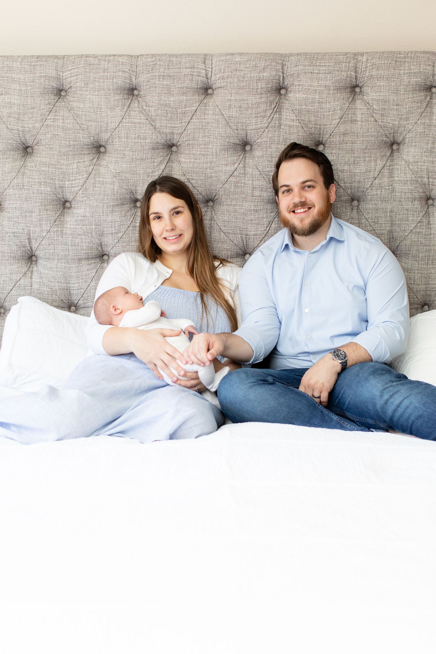 Family portrait with newborn baby against grey tufted headboard.