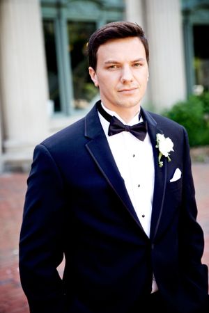 A formal portrait of a groom in a classic black tuxedo with bow tie and boutonniere.