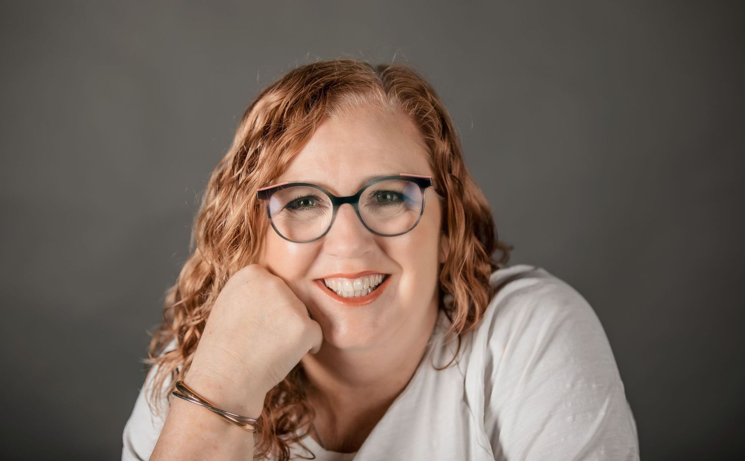 Professional headshot of someone with red curly hair wearing glasses and a white shirt against a gray backdrop.