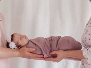 Newborn baby wearing mauve wrap and headband lying peacefully on supportive hands.