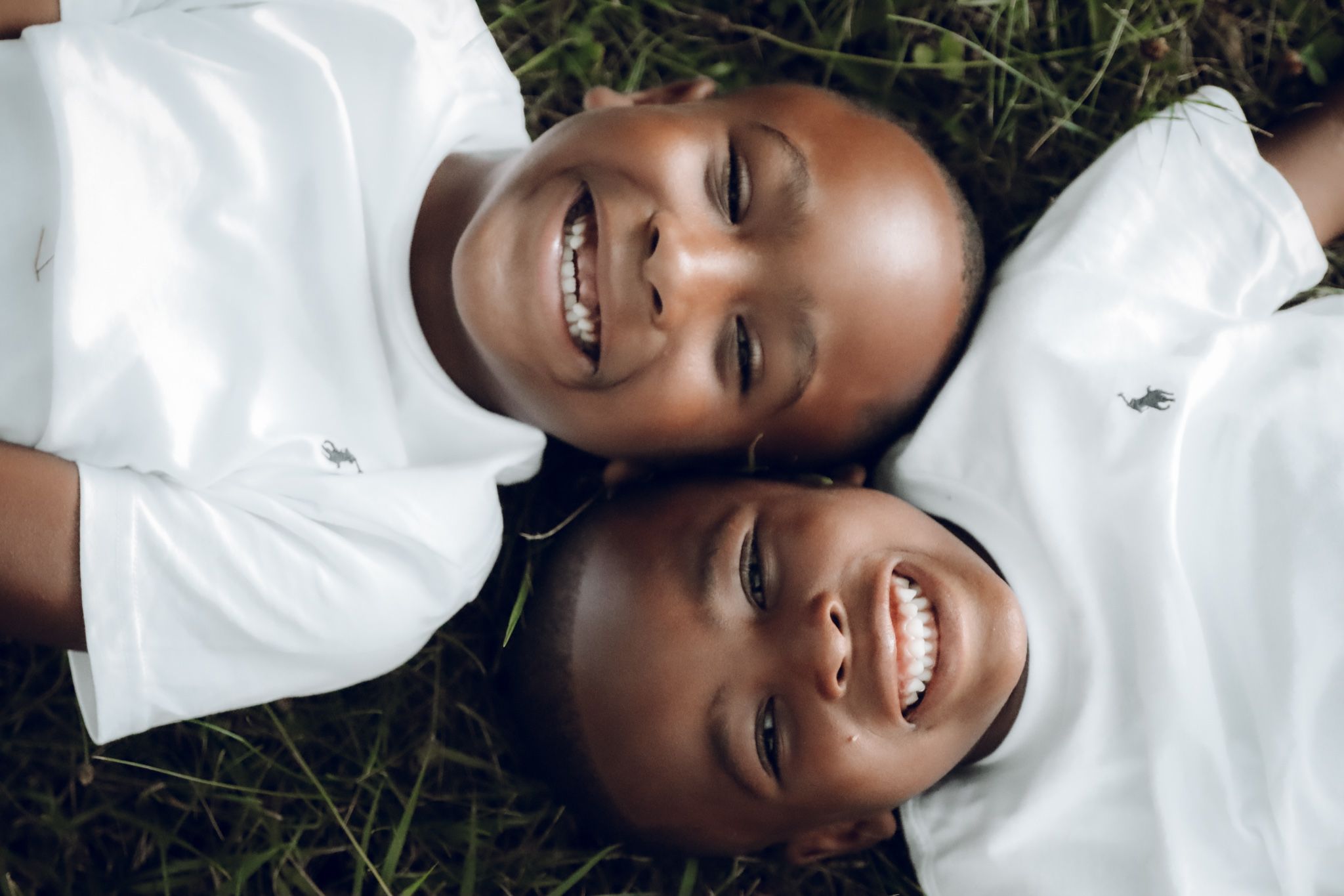 Two children in white shirts lying on grass smiling at camera.