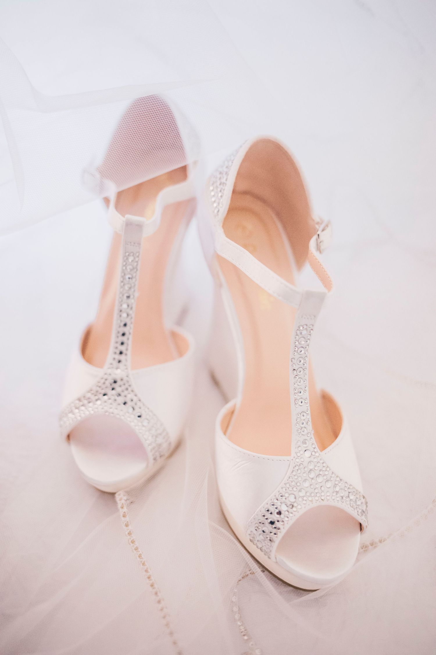 Elegant white bridal heels with crystal T-strap detail displayed on white surface.