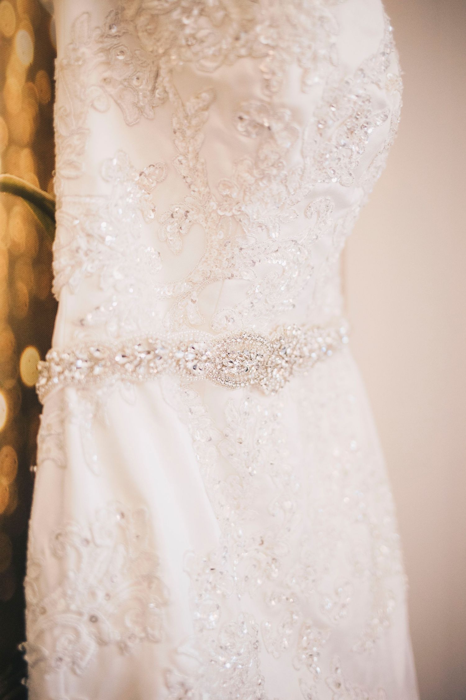 Close-up of beaded white wedding dress with rhinestone belt hanging on display.