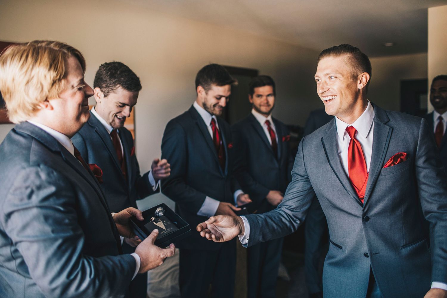 Groomsmen in gray suits with red ties laughing and shaking hands before a wedding ceremony.