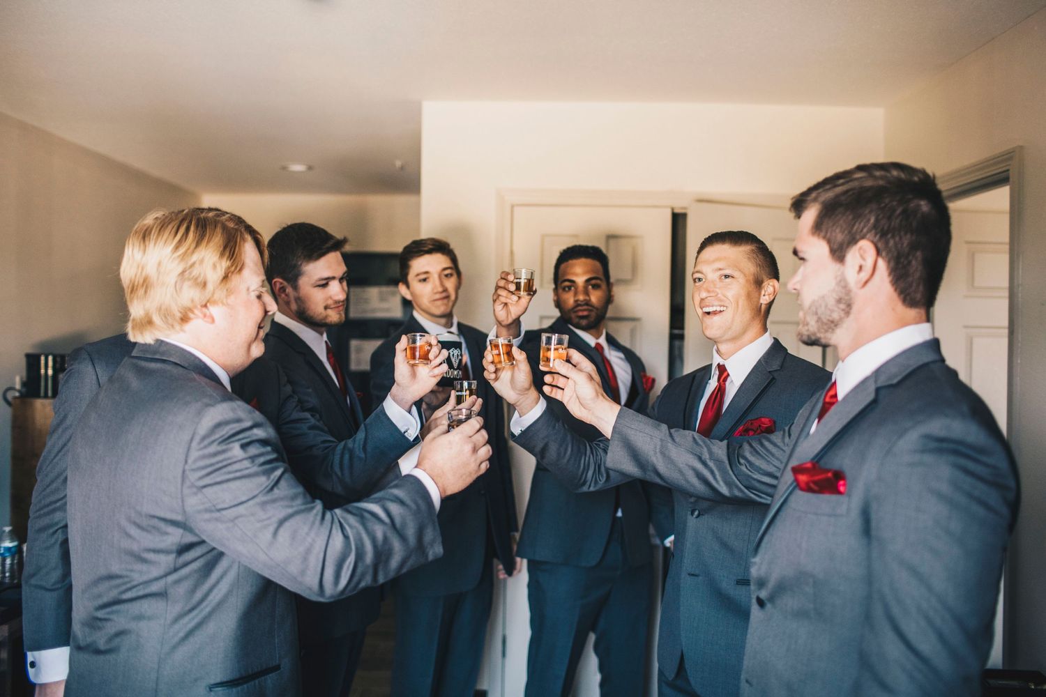 Groomsmen in gray suits toasting with drinks before a wedding celebration.