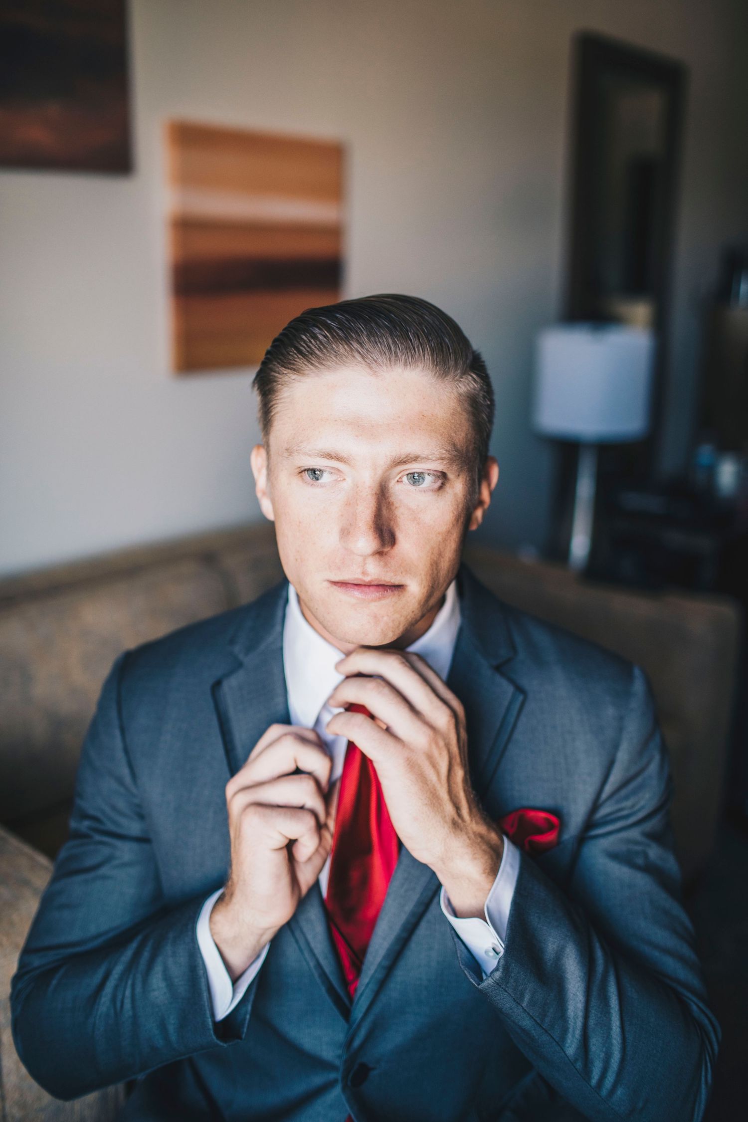 Groom in gray suit adjusting red tie while sitting in a hotel room before wedding.
